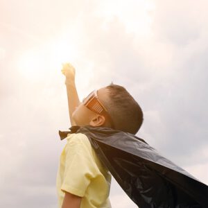 Young boy wearing a superhero cape and sun glasses reaching to the sun